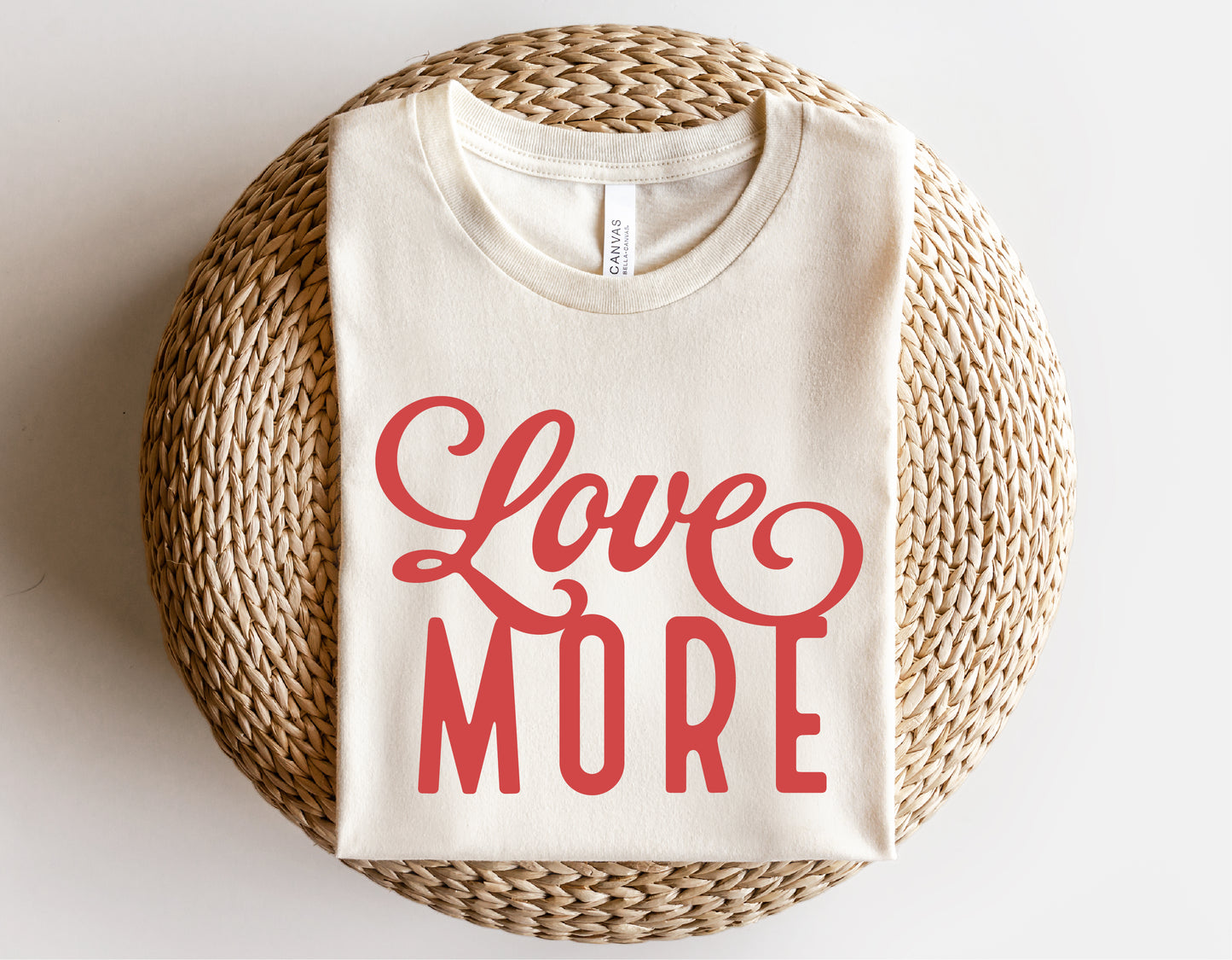 White t-shirt with "Love More" text in red, neatly folded on a woven round mat, showcasing Valentine's Day clothing design.