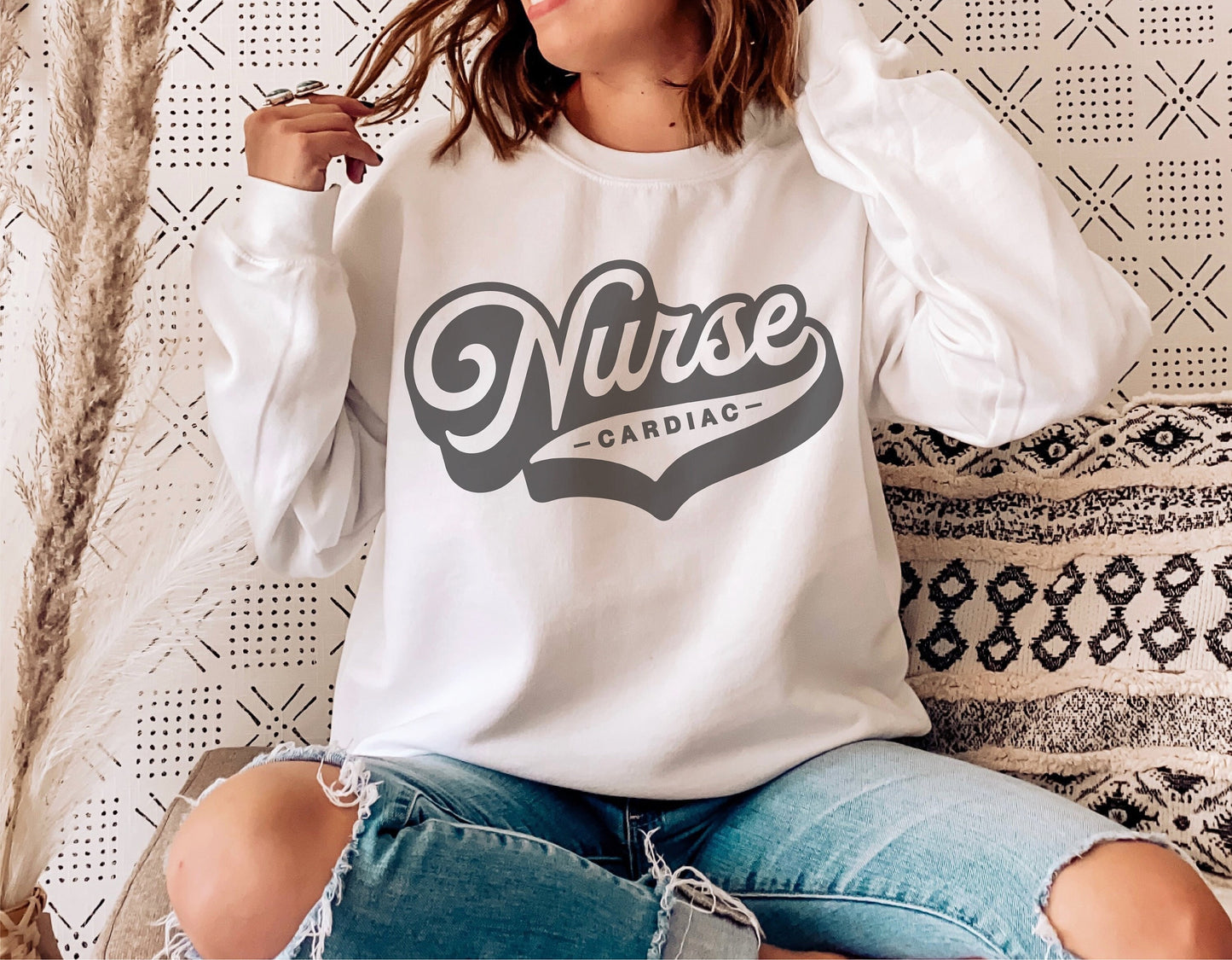 Woman wearing white sweatshirt with 'Cardiac Nurse' retro design, sitting casually against decorative wall and pillows.