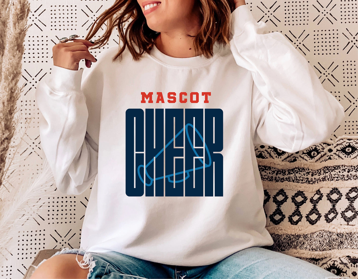 Woman wearing 'Mascot Cheer' sweatshirt with graphic design, sitting on a couch.