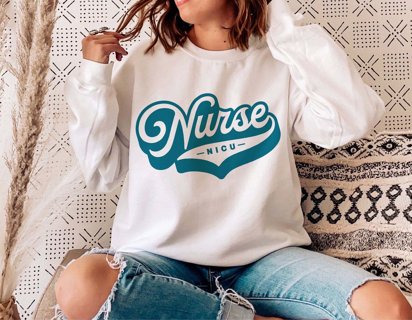 Woman wearing white sweatshirt with teal 'NICU Nurse' design, sitting on textured cushion. Ideal for nurse-themed SVG projects.