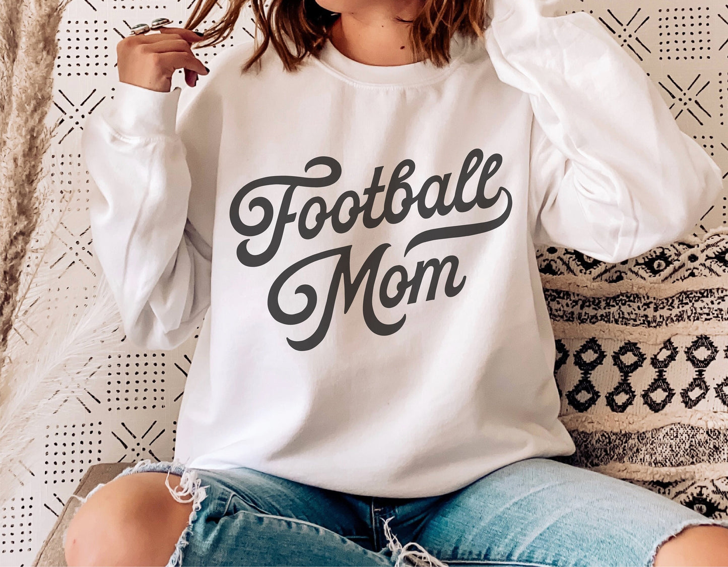 Woman wearing a white sweatshirt with 'Football Mom' text, sitting on a patterned couch, casual and vintage style.