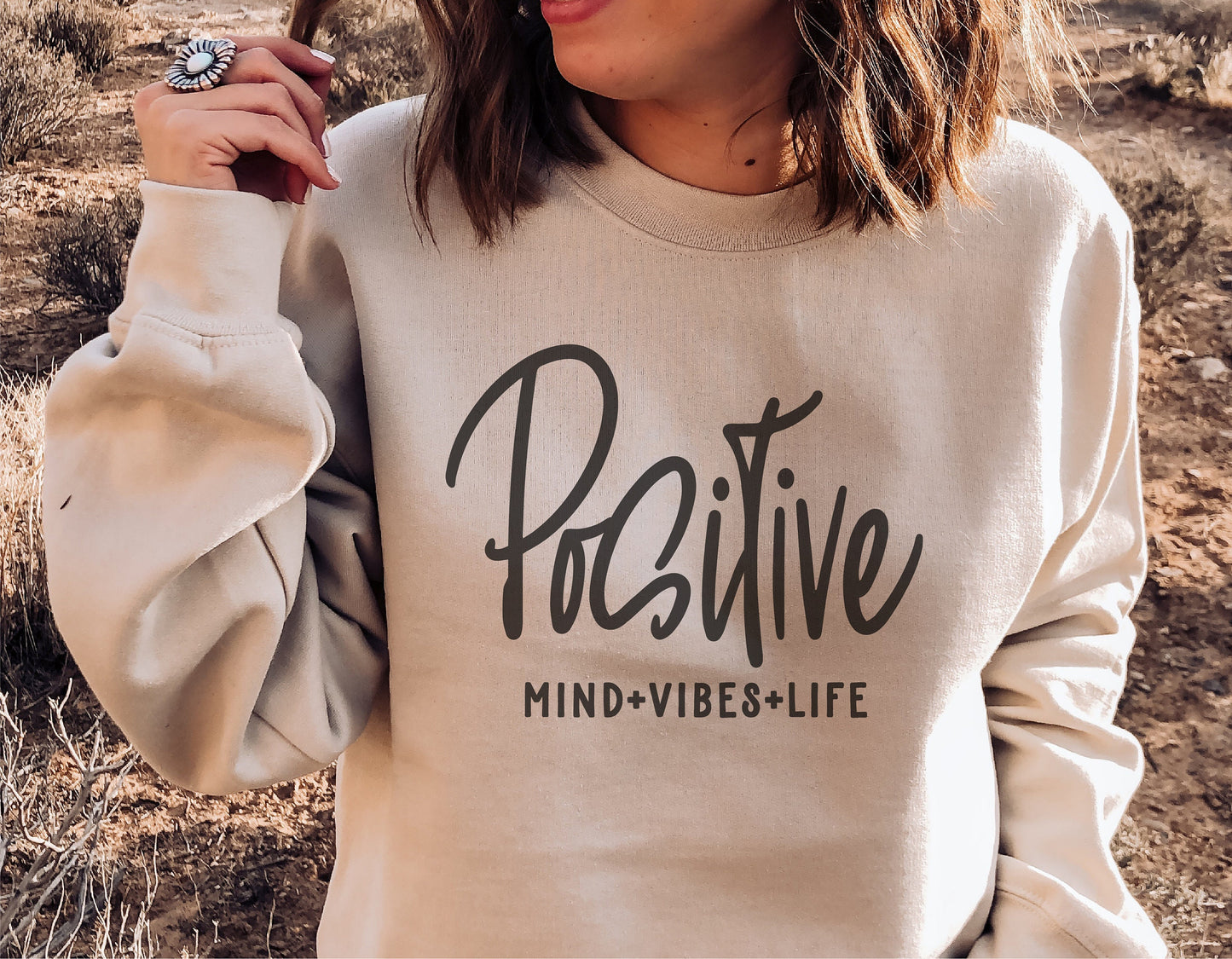 Woman wearing a beige sweatshirt with Positive Mind Positive Vibes Positive Life graphic in outdoor setting