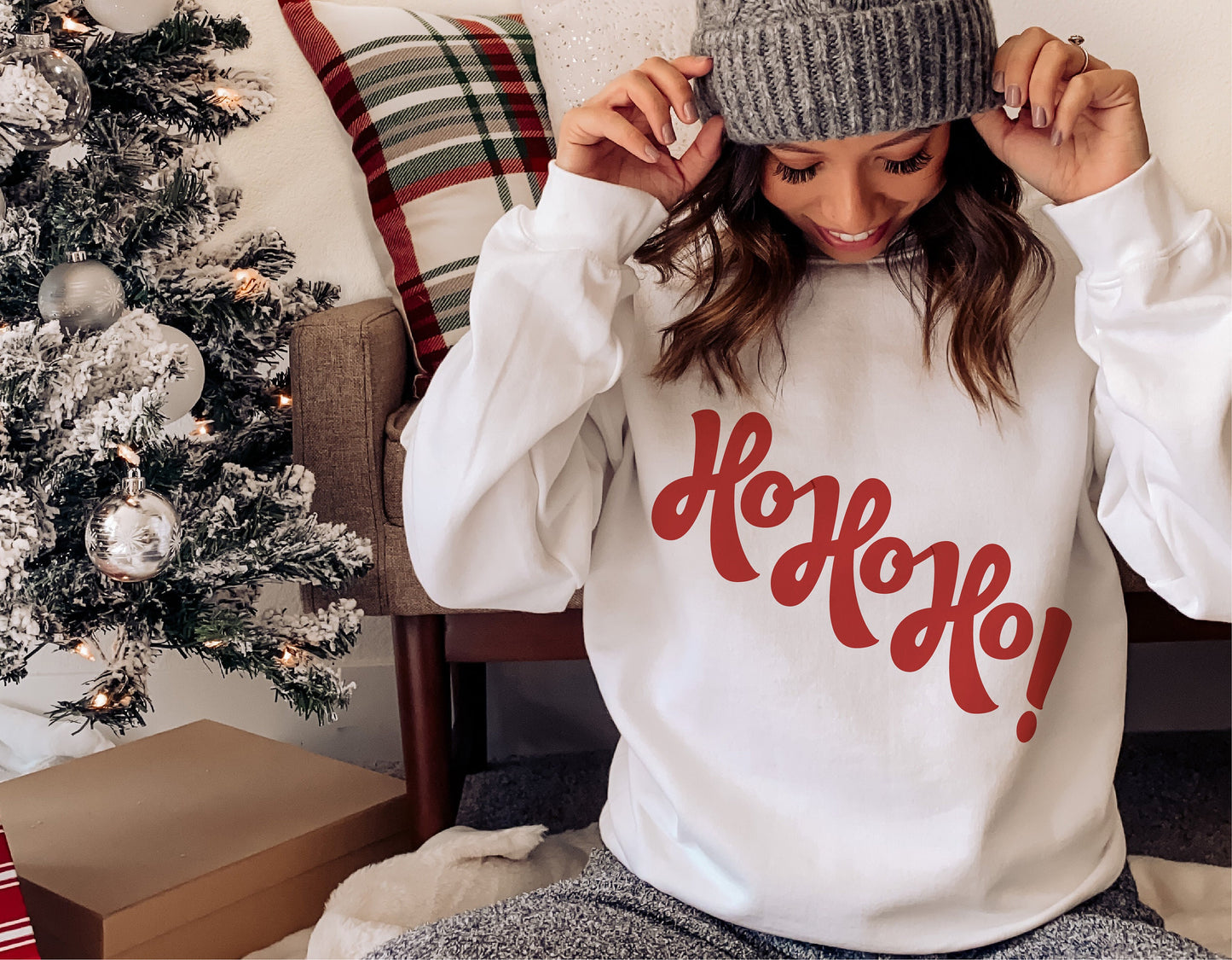 Woman wearing a funny 'Ho Ho Ho' Christmas sweater with Santa design, sitting by a decorated tree, ideal for winter holiday cheer.