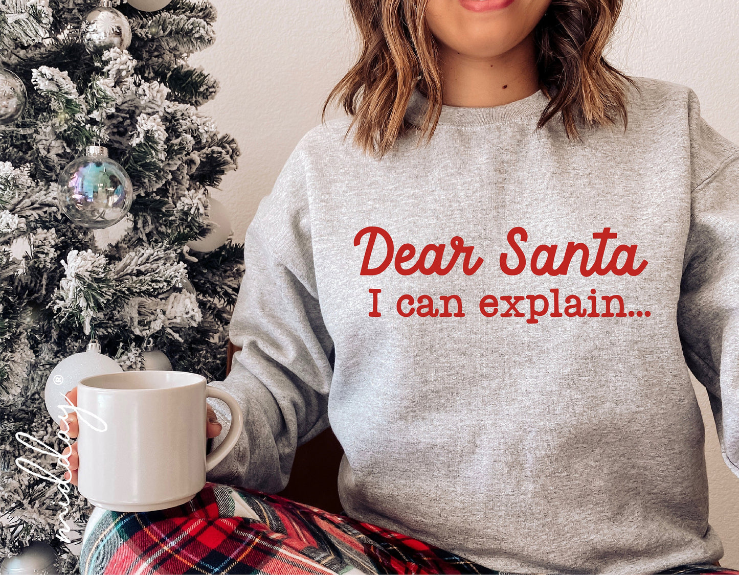 Woman wearing 'Dear Santa I Can Explain' sweatshirt, holding a mug by a Christmas tree, festive and funny holiday scene.
