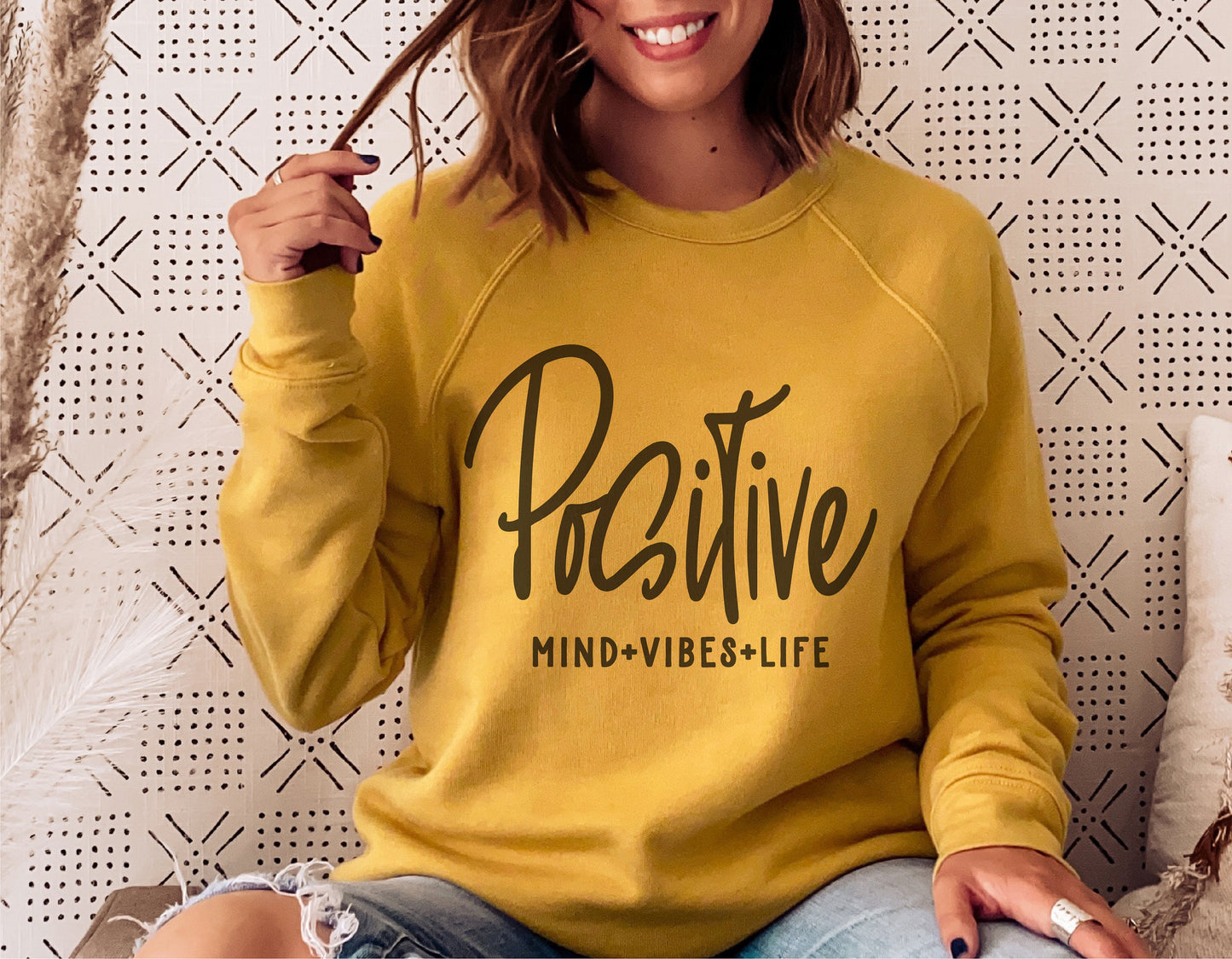 Woman wearing yellow sweatshirt with 'Positive Mind Positive Vibes Positive Life' print, sitting in cozy setting.
