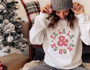 Woman wearing a Fa La La Ho Ho Ho white Christmas sweater, sitting by a decorated tree and gifts, cozy winter holiday scene.