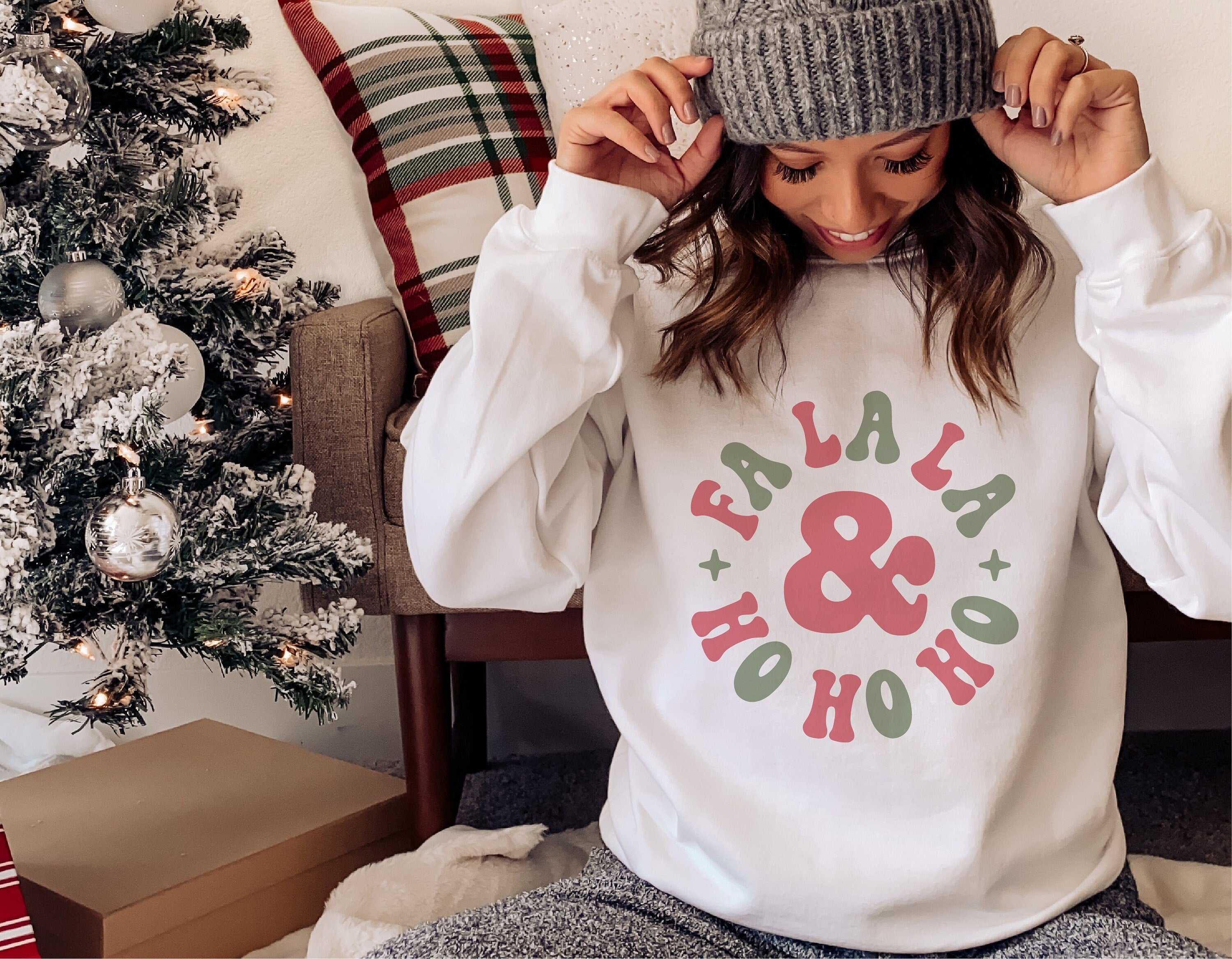 Woman wearing a Fa La La Ho Ho Ho white Christmas sweater, sitting by a decorated tree and gifts, cozy winter holiday scene.
