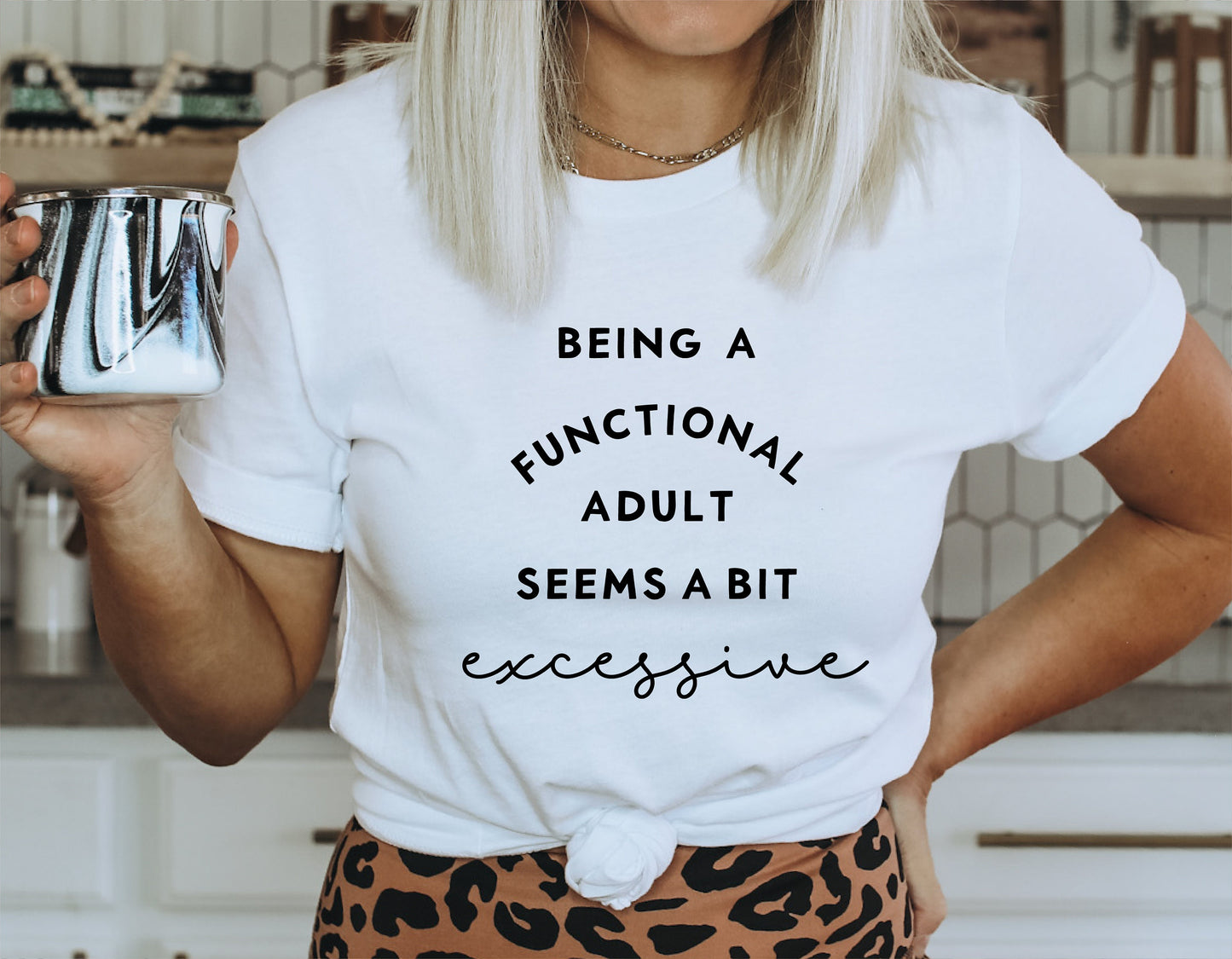 Woman wearing a white t-shirt with 'Being a Functional Adult Seems a Bit Excessive' text, holding a coffee mug in modern kitchen. Sarcastic design.