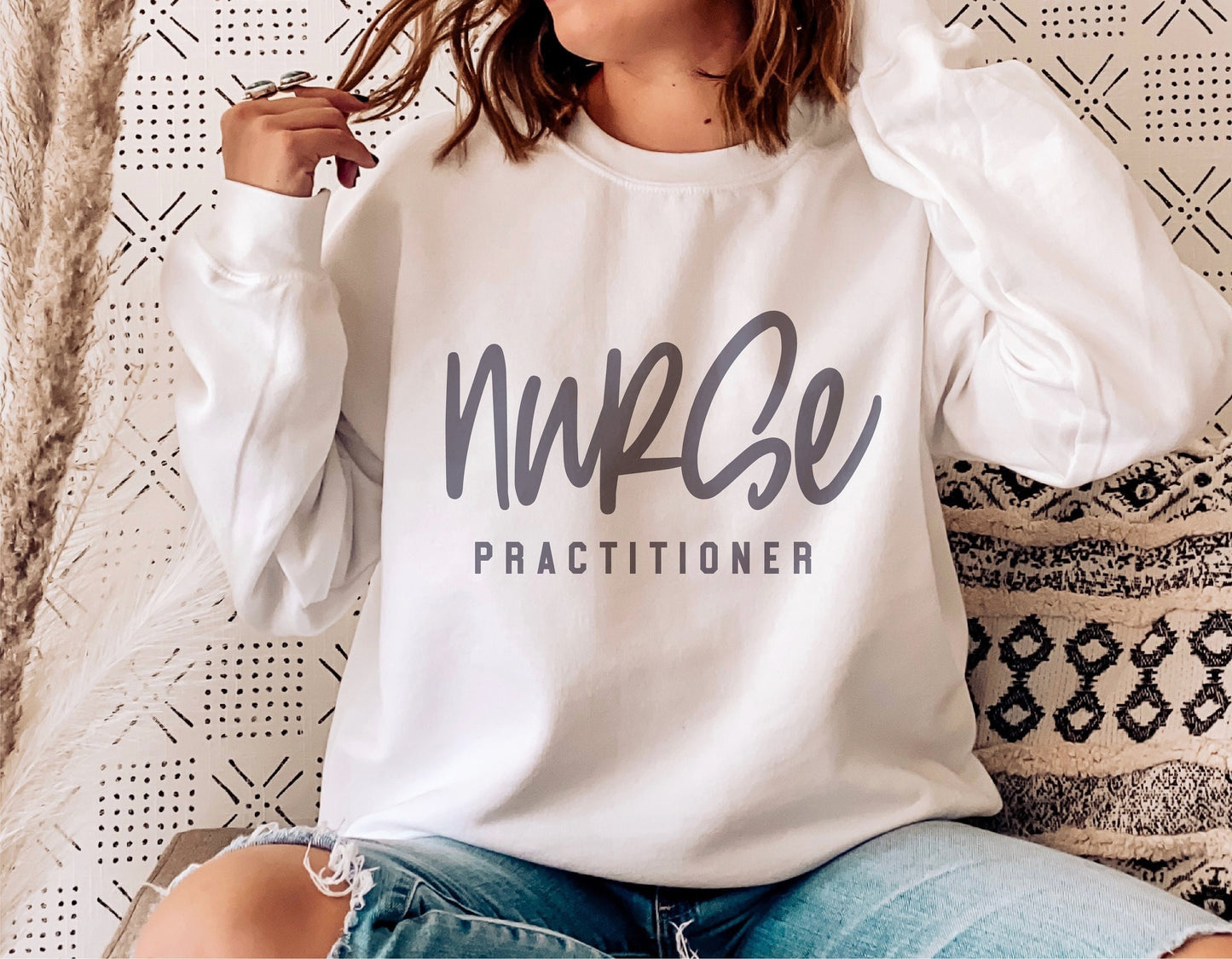 Woman wearing a white 'Nurse Practitioner' sweatshirt, sitting on a patterned cushion, perfect for nurse appreciation gifts.