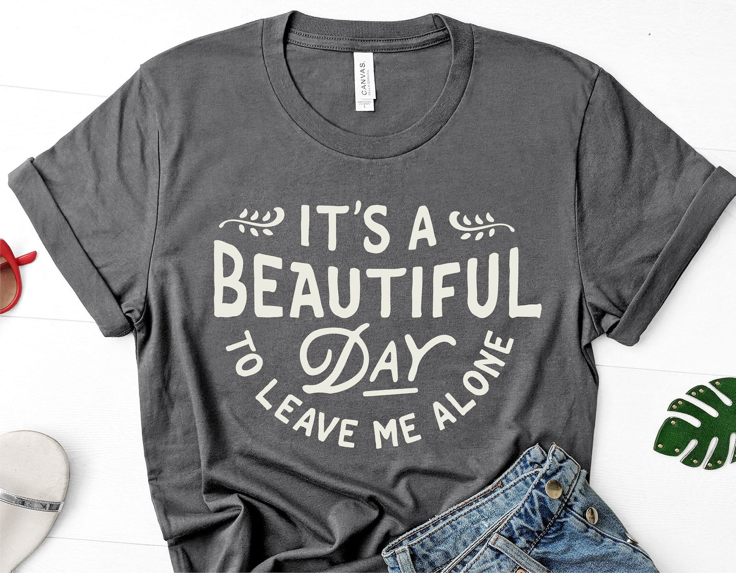 Gray t-shirt with 'It's a Beautiful Day to Leave Me Alone' text, displayed with casual denim and summer accessories.