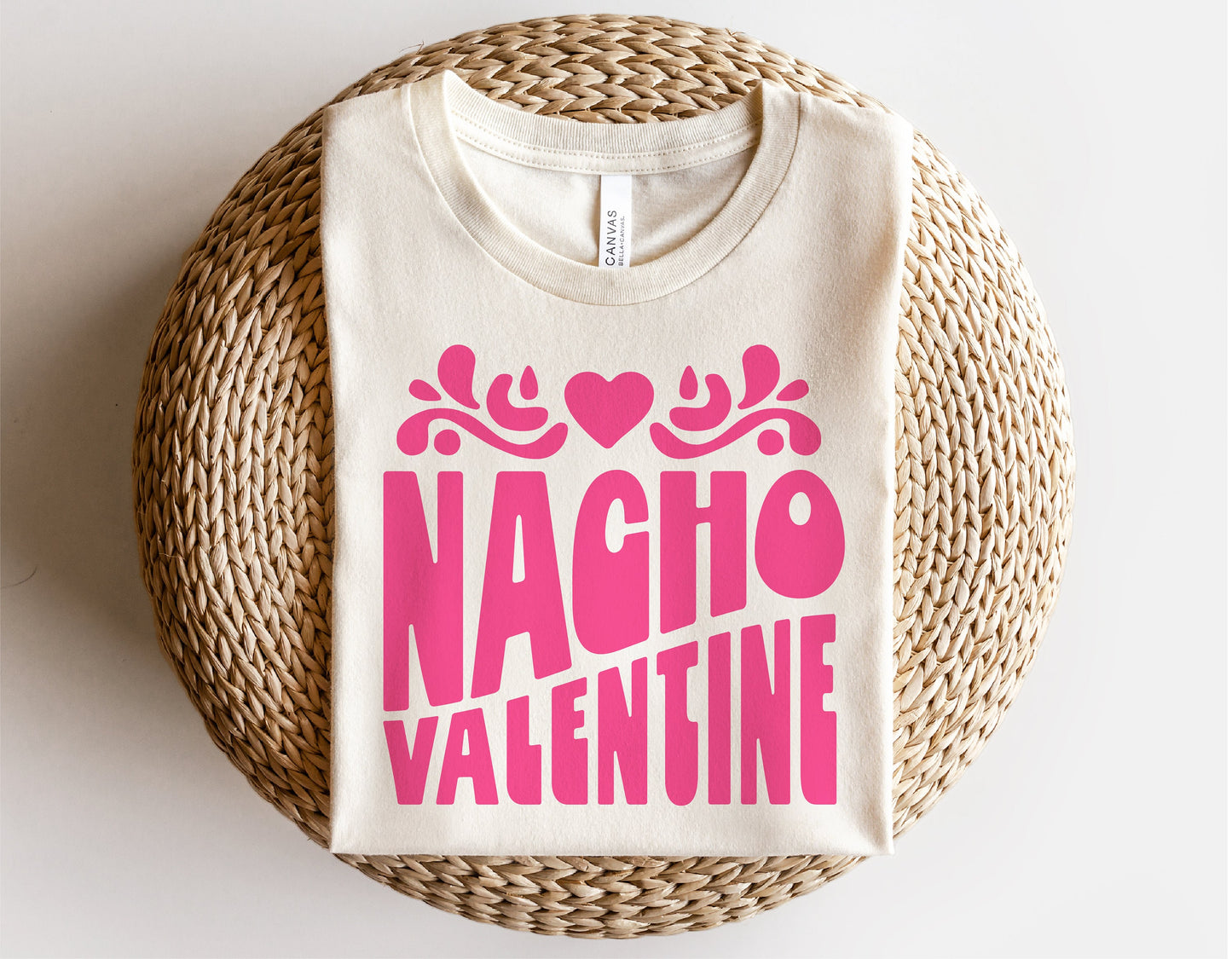 T-shirt with "Nacho Valentine" pink text and heart design, folded on a woven surface, perfect for humorous Valentine's Day gifting.