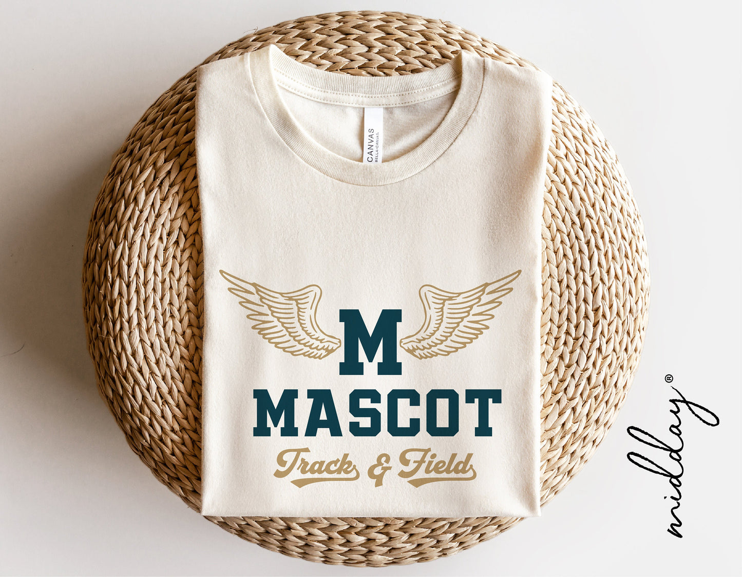 Track and field logo shirt with 'M Mascot' and wing design, folded on a woven basket, perfect for sports team gifts and Cricut projects.