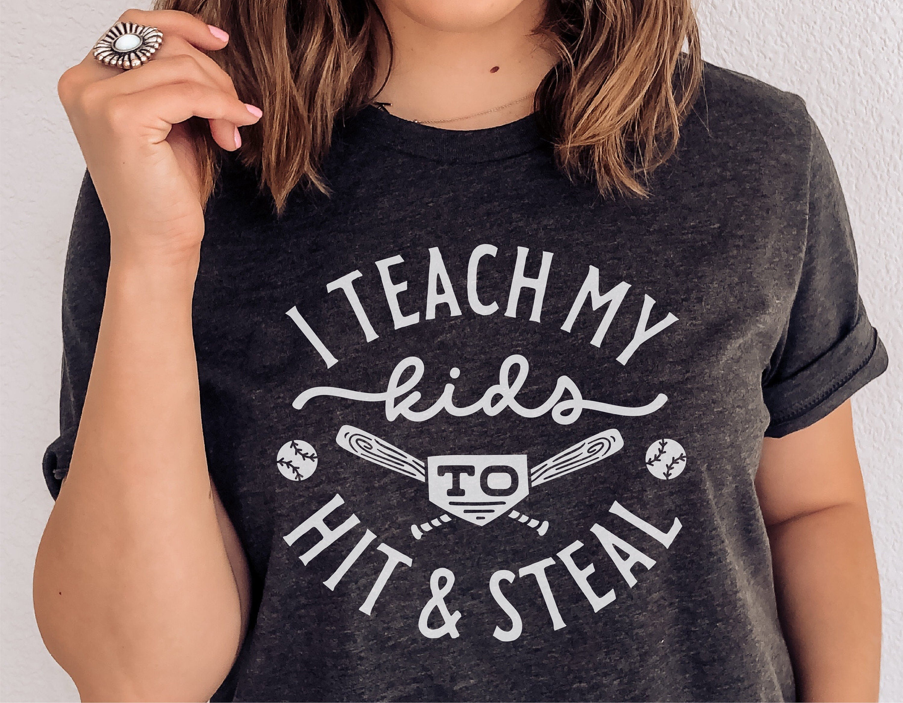 Woman wearing a funny baseball shirt, "I Teach My Kids to Hit & Steal," featuring baseball graphics and bats, perfect for game day.