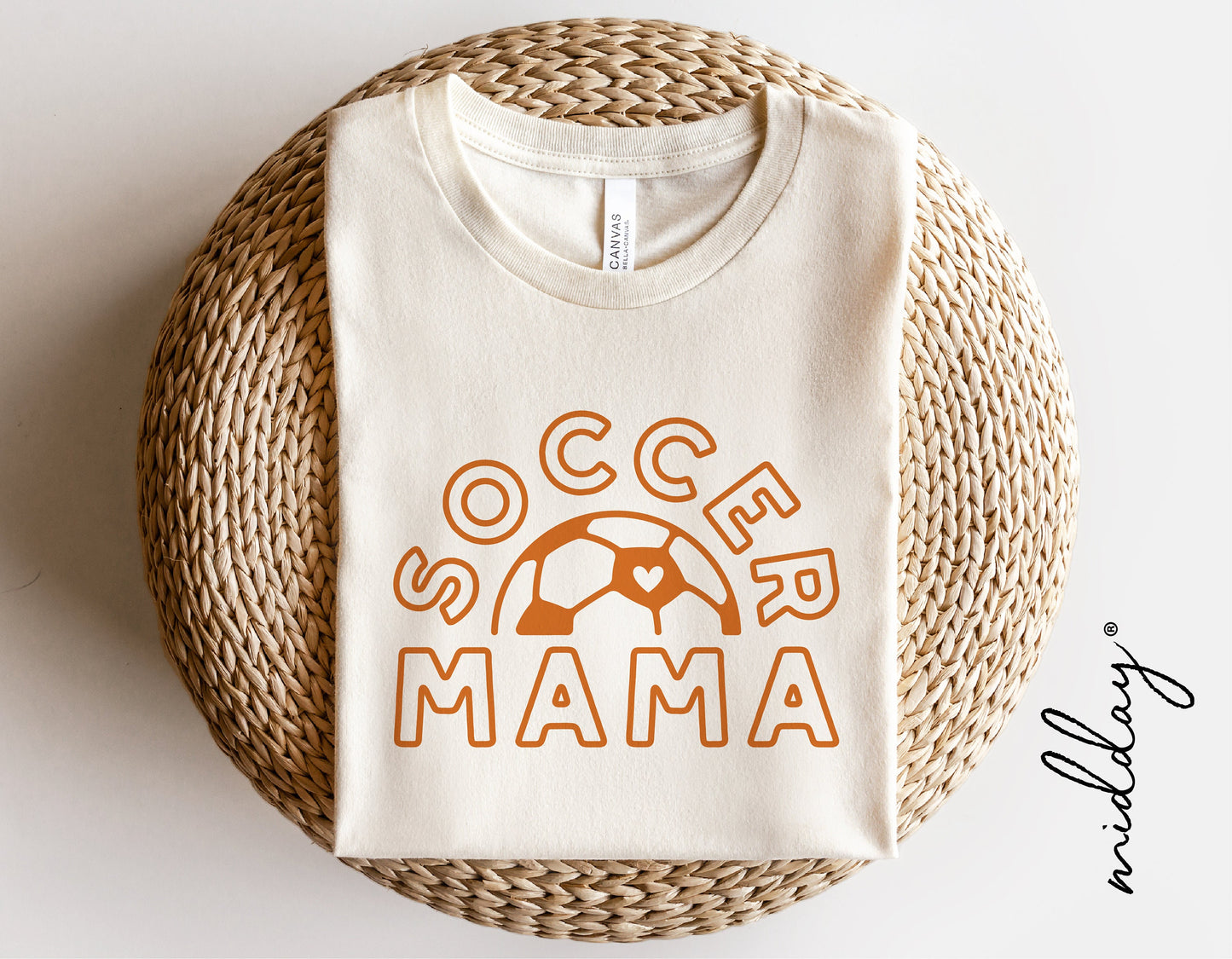 White 'Soccer Mama' shirt with orange text and soccer ball graphic, folded on a woven mat, perfect for soccer mom gifts and crafts.