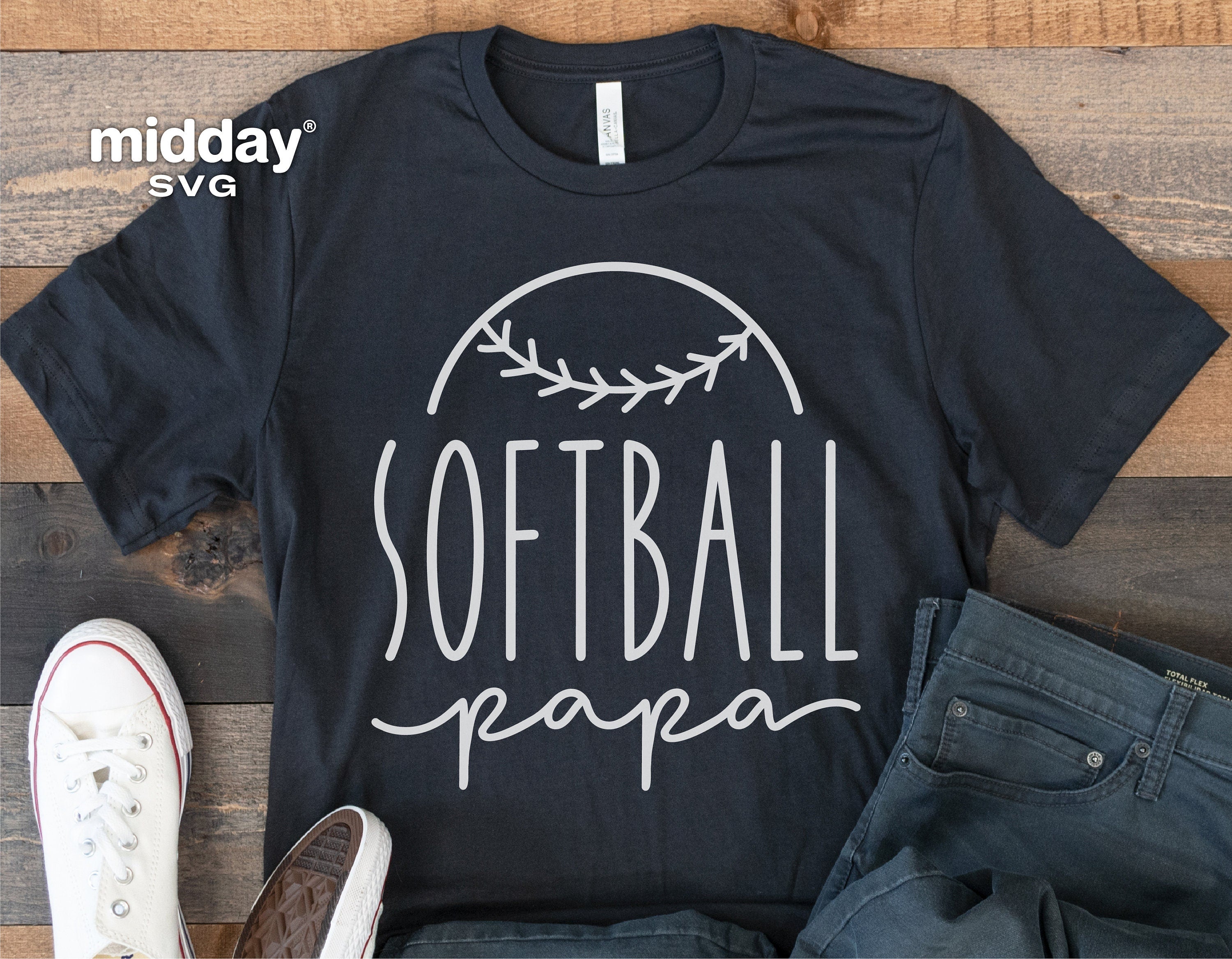 Black t-shirt with 'Softball Papa' design, surrounded by casual footwear and jeans on a wooden floor.