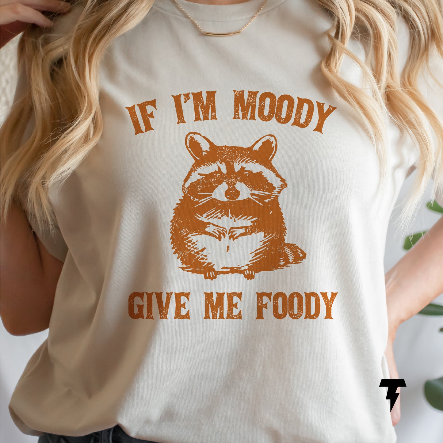 Woman wearing a beige T-shirt with a cute raccoon illustration and text 'If I'm Moody Give Me Foody,' perfect for food lovers.