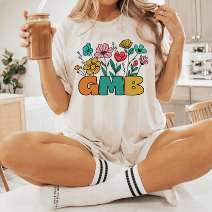 Unisex GMB t-shirt with vibrant floral design, worn by a person in a relaxed setting with a drink, showcasing comfy style.