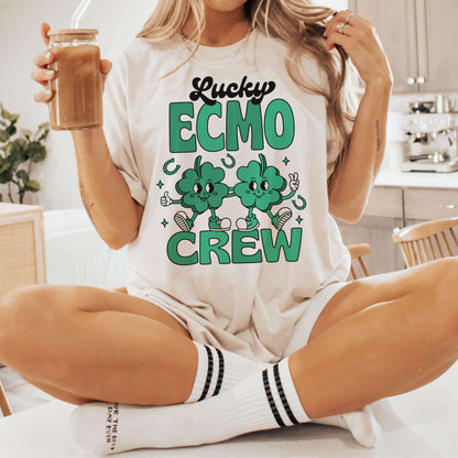 Woman wearing Lucky ECMO Crew St. Patrick's Day shirt with shamrock design, sipping drink on kitchen counter.