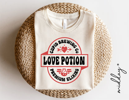 Cupid Brewing Co Love Potion t-shirt folded on wicker basket, featuring premium elixirs design with heart and wings.