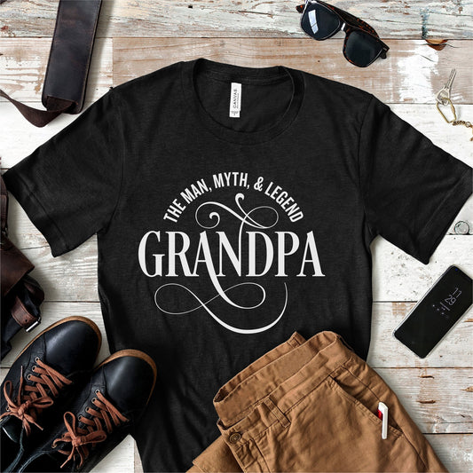 Black t-shirt with 'The Man, Myth, & Legend Grandpa' text, surrounded by men's accessories on a wooden surface.