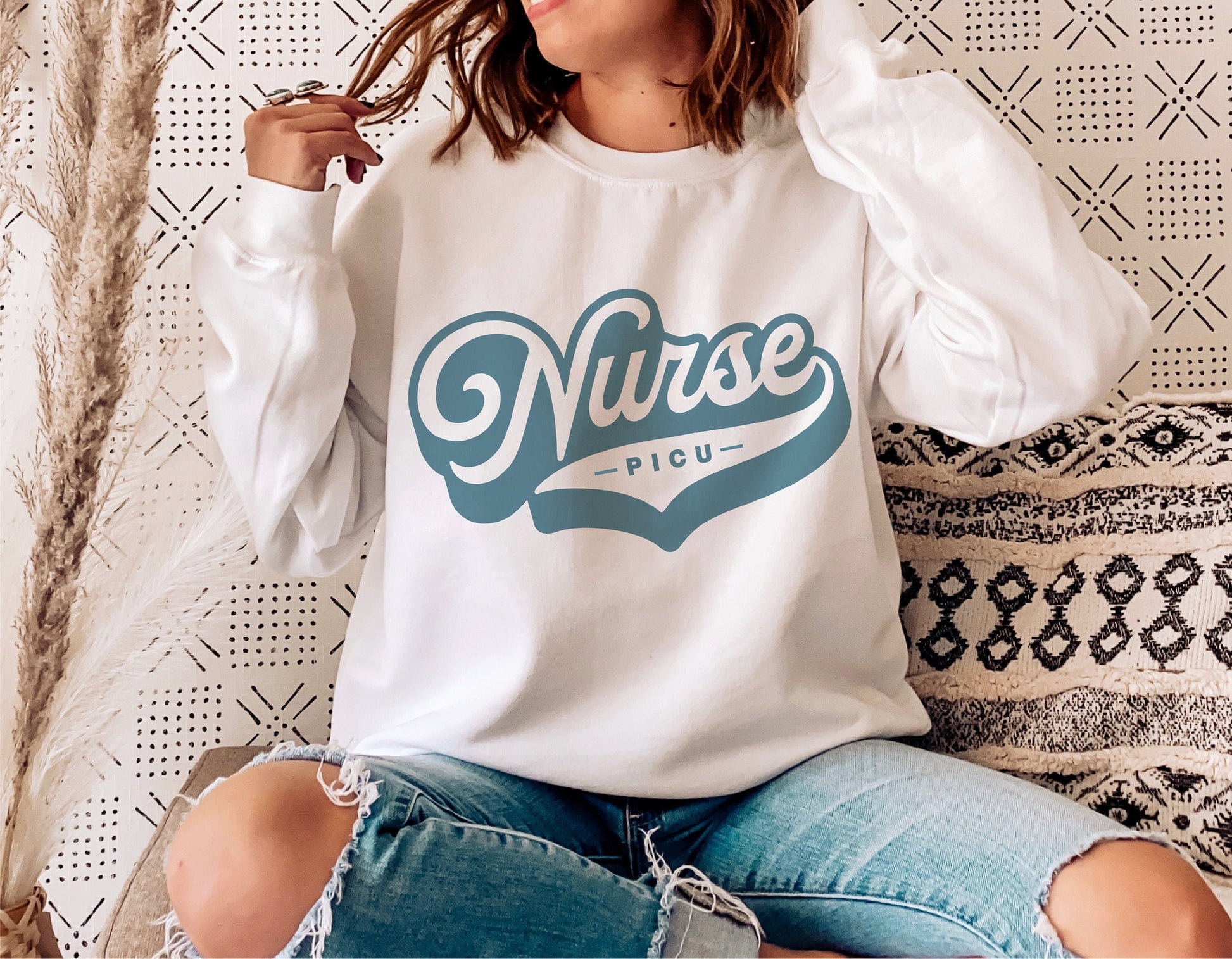 PICU Nurse logo on white sweatshirt, worn by person sitting on couch decorated with modern patterns; cozy, retro, and trendy design.