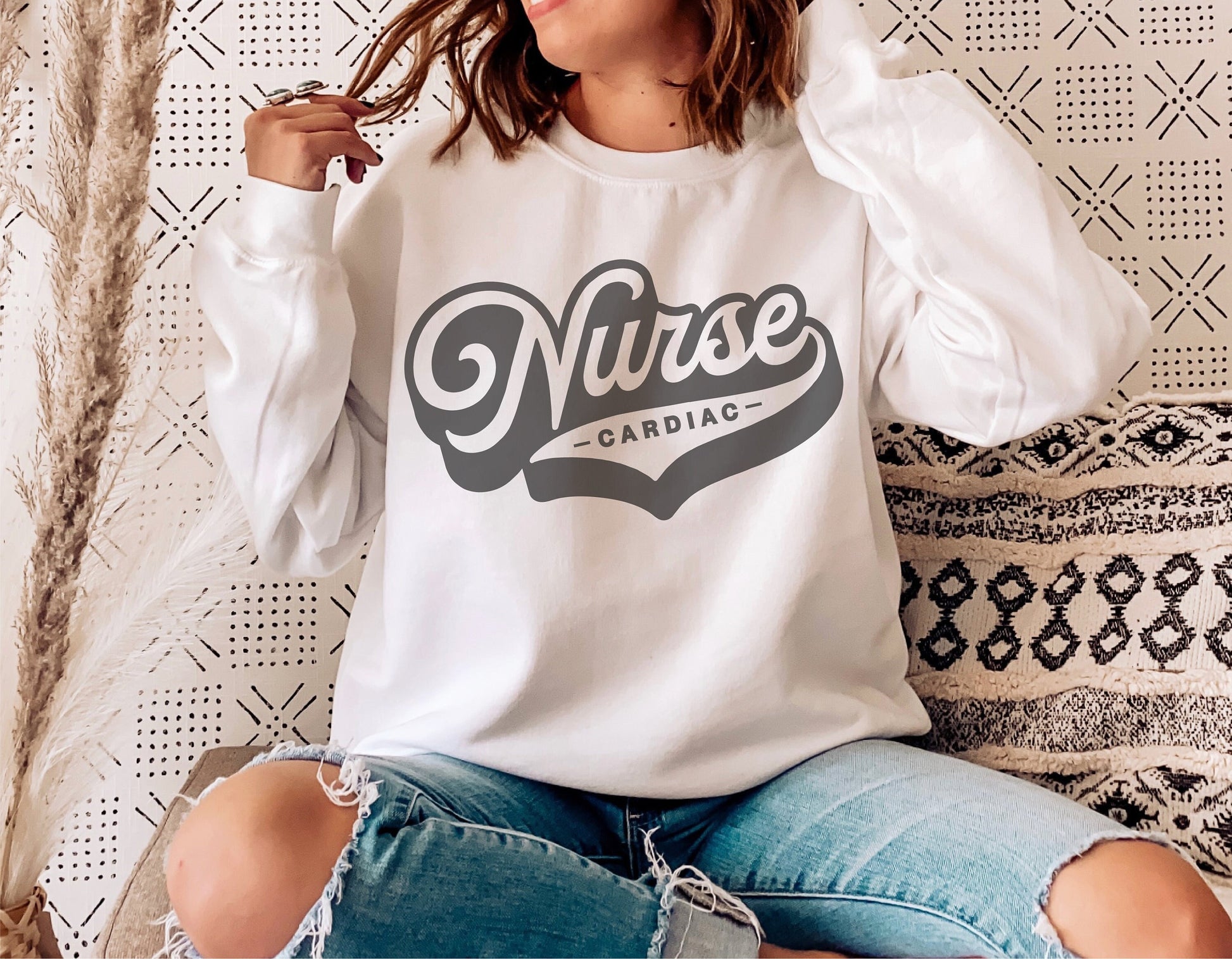 Woman wearing white sweatshirt with 'Cardiac Nurse' retro design, sitting casually against decorative wall and pillows.