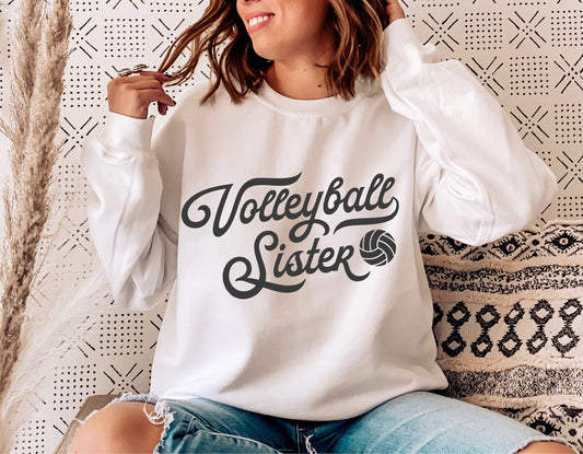 Woman wearing a white sweatshirt with 'Volleyball Sister' design, sitting casually with a vintage pattern background.