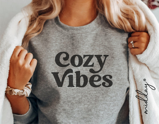 Woman wearing a grey sweater with 'Cozy Vibes' text, wrapped in a white blanket, showcasing cozy comfort style.
