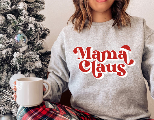 Woman wearing 'Mama Claus' Christmas sweater sitting by decorated tree with mug