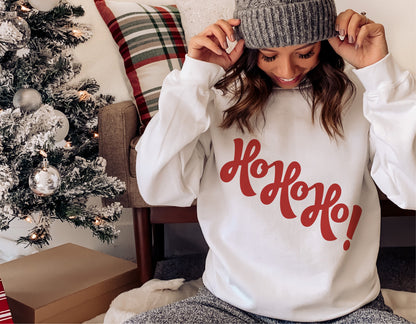 Woman wearing a funny 'Ho Ho Ho' Christmas sweater with Santa design, sitting by a decorated tree, ideal for winter holiday cheer.