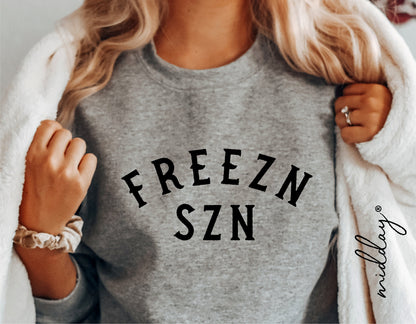 Woman wearing a gray sweatshirt with black 'FREEZN SZN' text, cozy holiday fashion.