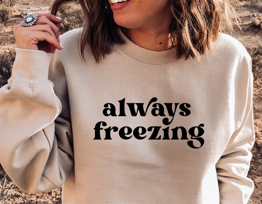 Woman wearing a beige sweatshirt with the text 'Always Freezing', ideal for winter fashion and holiday gifts.