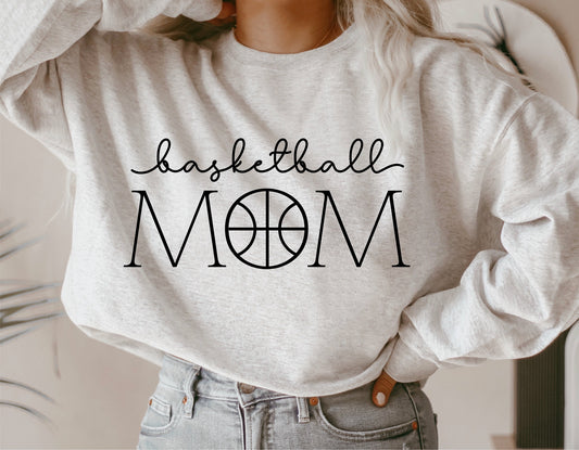 Woman wearing a light gray sweatshirt with 'Basketball Mom' design featuring a basketball graphic in the letter 'O'.