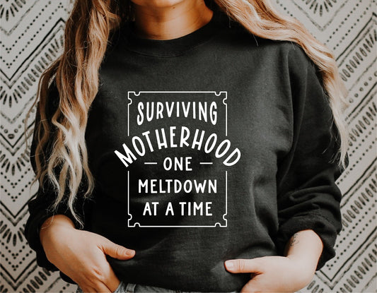 Woman wearing black sweatshirt with 'Surviving Motherhood One Meltdown at a Time' text, funny mom svg design for Cricut.