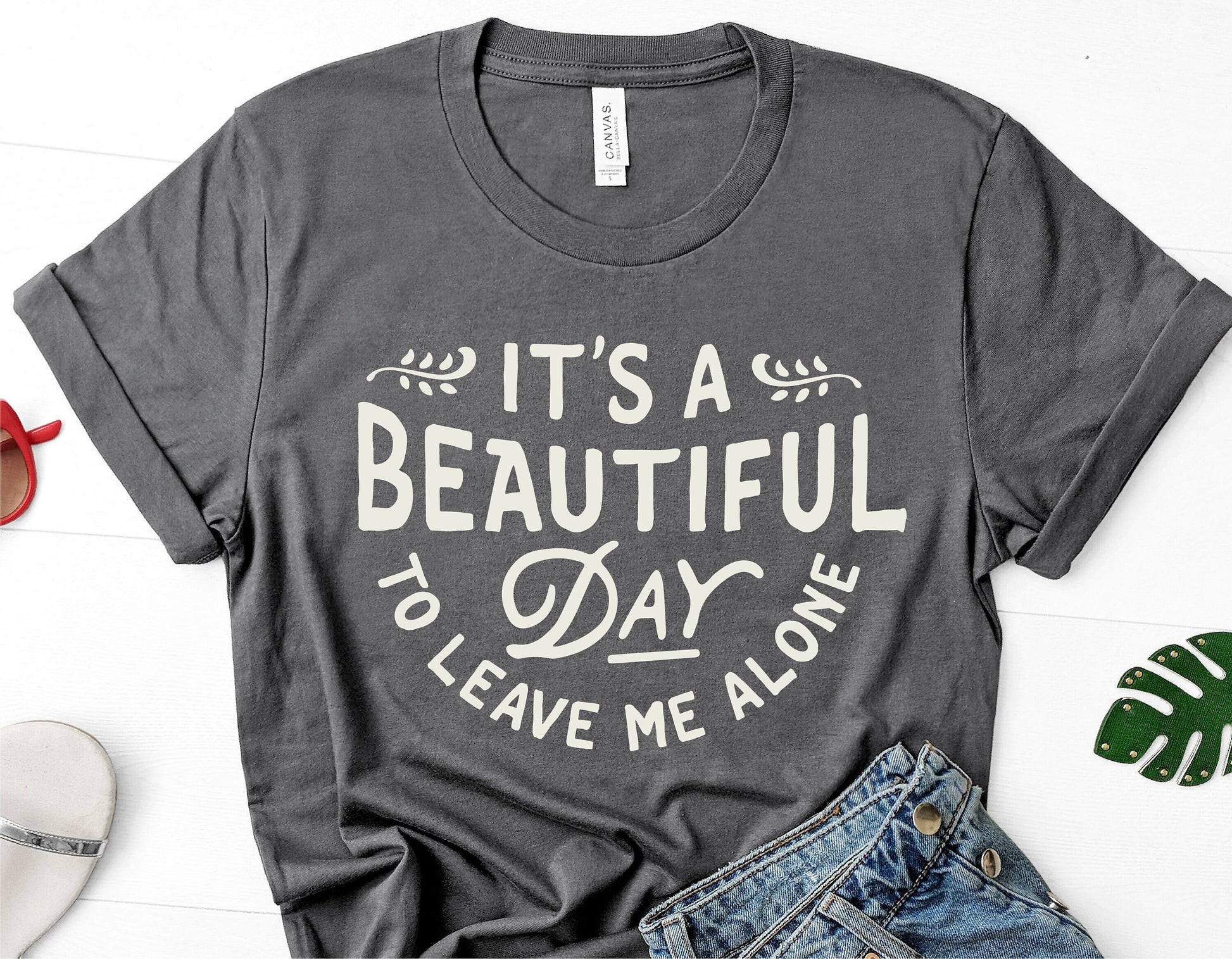 Gray t-shirt with 'It's a Beautiful Day to Leave Me Alone' text, displayed with casual denim and summer accessories.