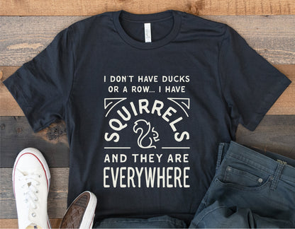 Black T-shirt with "I Have Squirrels Everywhere" funny design on wood background, paired with jeans and white sneakers.