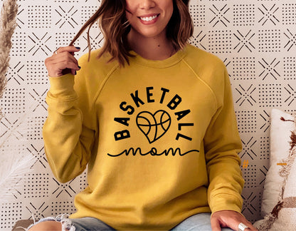 Woman wearing a yellow sweatshirt featuring a 'Basketball Mom' design with a heart-shaped basketball graphic, sitting on a couch.