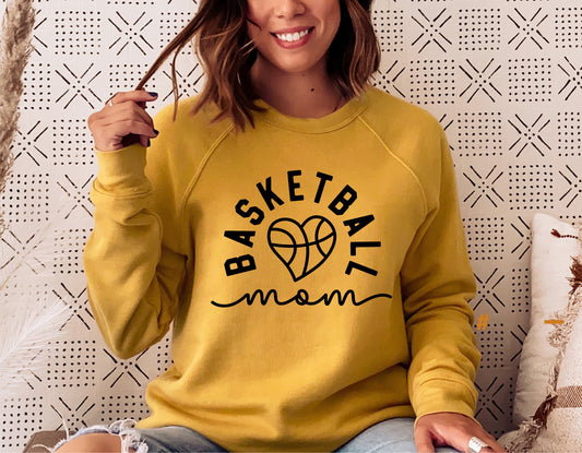Woman wearing a yellow sweatshirt featuring a 'Basketball Mom' design with a heart-shaped basketball graphic, sitting on a couch.