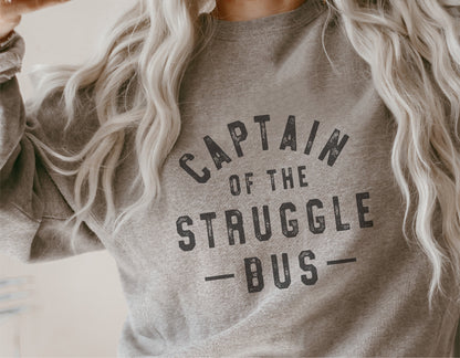 Woman wearing a grey 'Captain of the Struggle Bus' sweatshirt with wavy blonde hair.