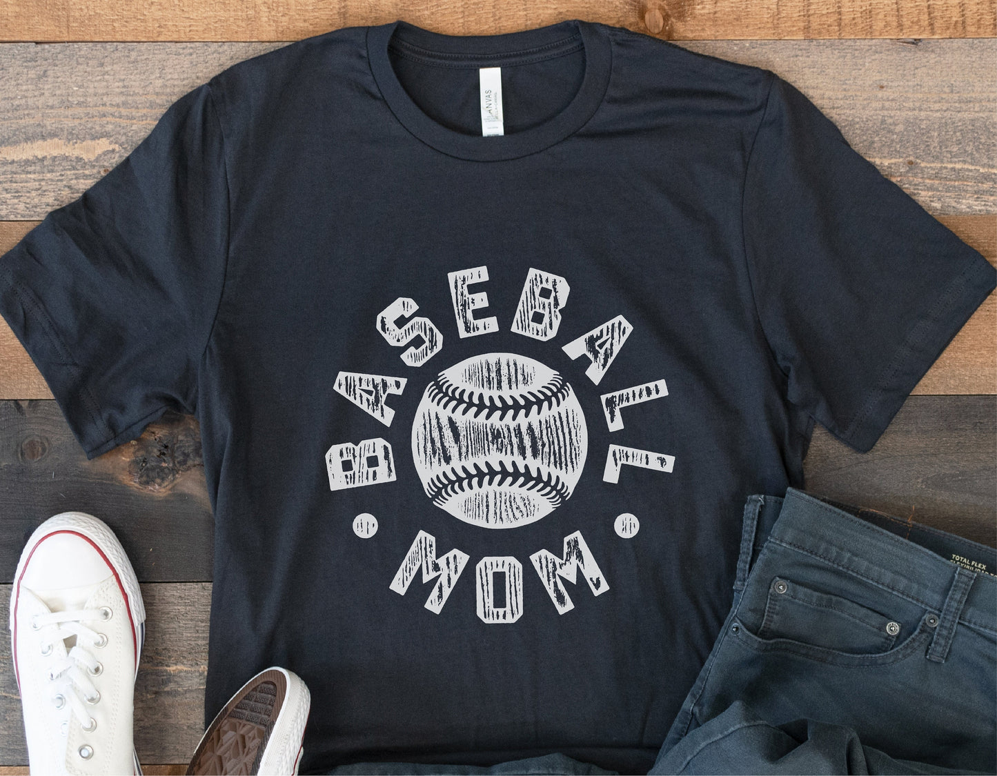 Black t-shirt with "Baseball Mom" graphic design next to white sneakers and jeans on wooden floor.