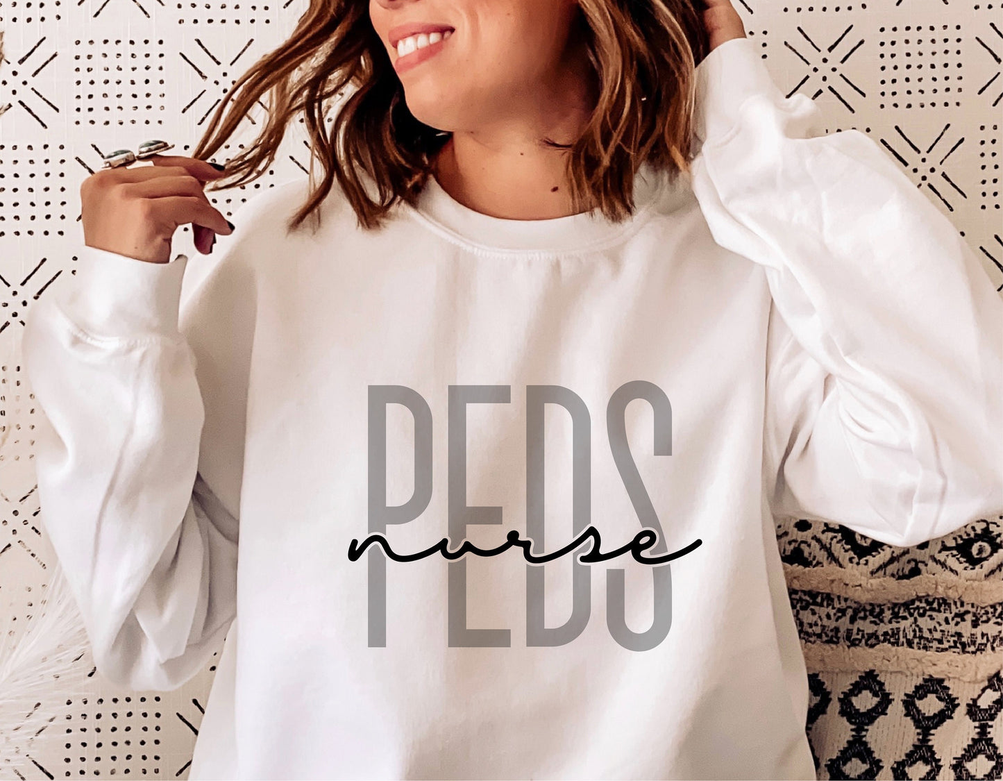 White sweatshirt with 'PEDS nurse' design, ideal for pediatric nurses, shown in modern home setting.
