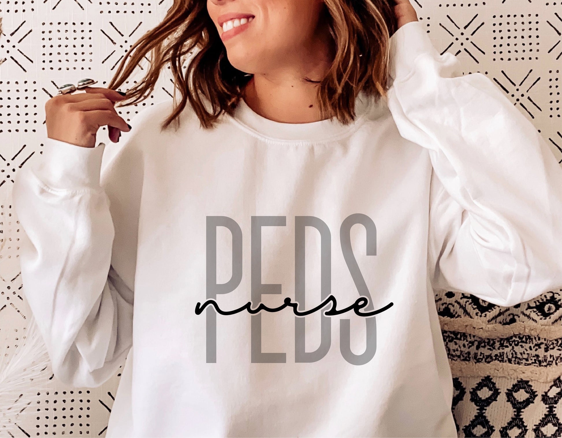 White sweatshirt with 'PEDS nurse' design, ideal for pediatric nurses, shown in modern home setting.