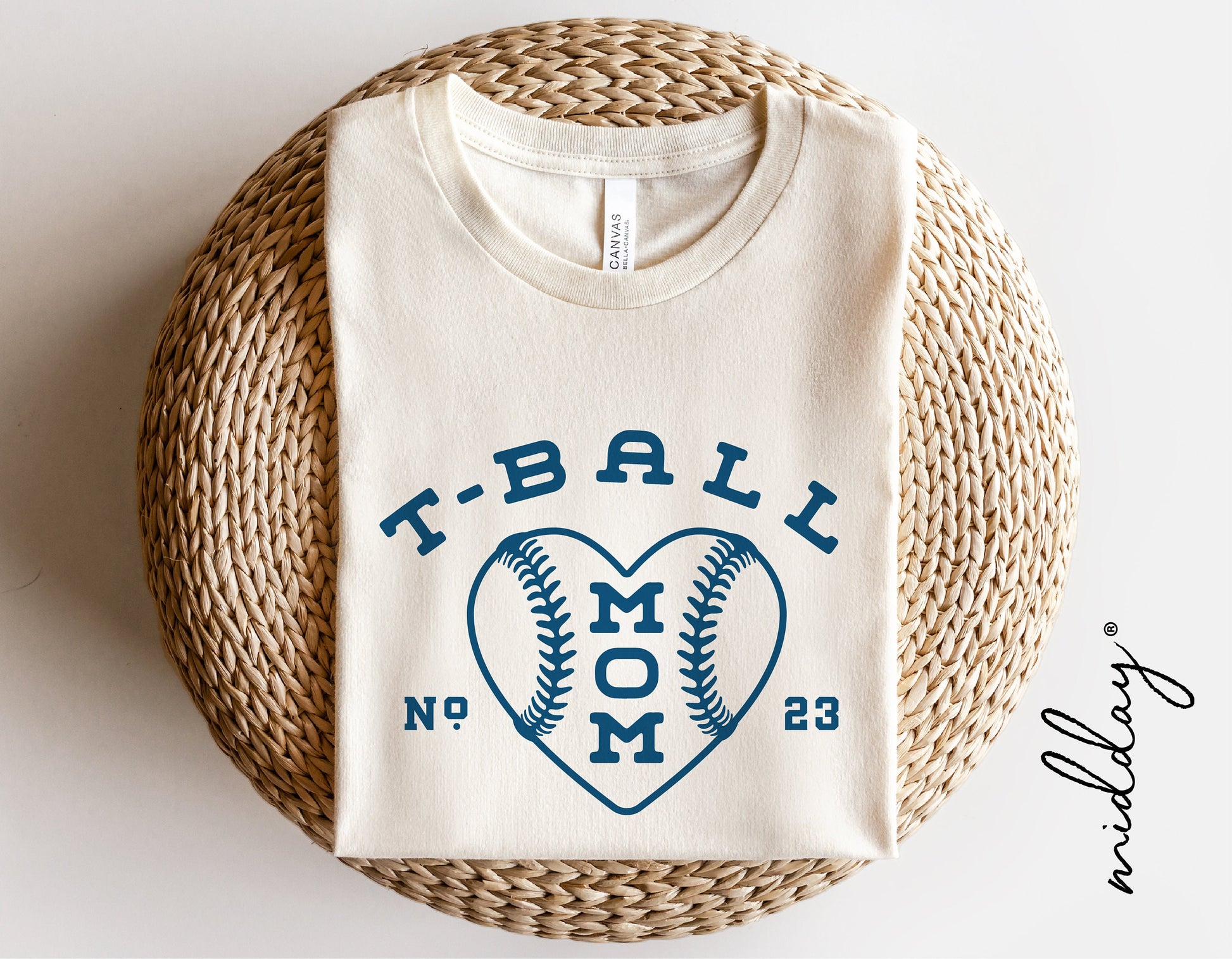 White T-shirt with "T-Ball Mom" design featuring a heart-shaped baseball graphic, displayed on a wicker basket.