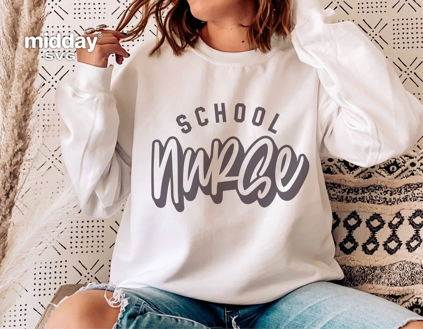 Person wearing a white sweatshirt with 'School Nurse' design in gray, sitting against a patterned background, showcasing casual style.