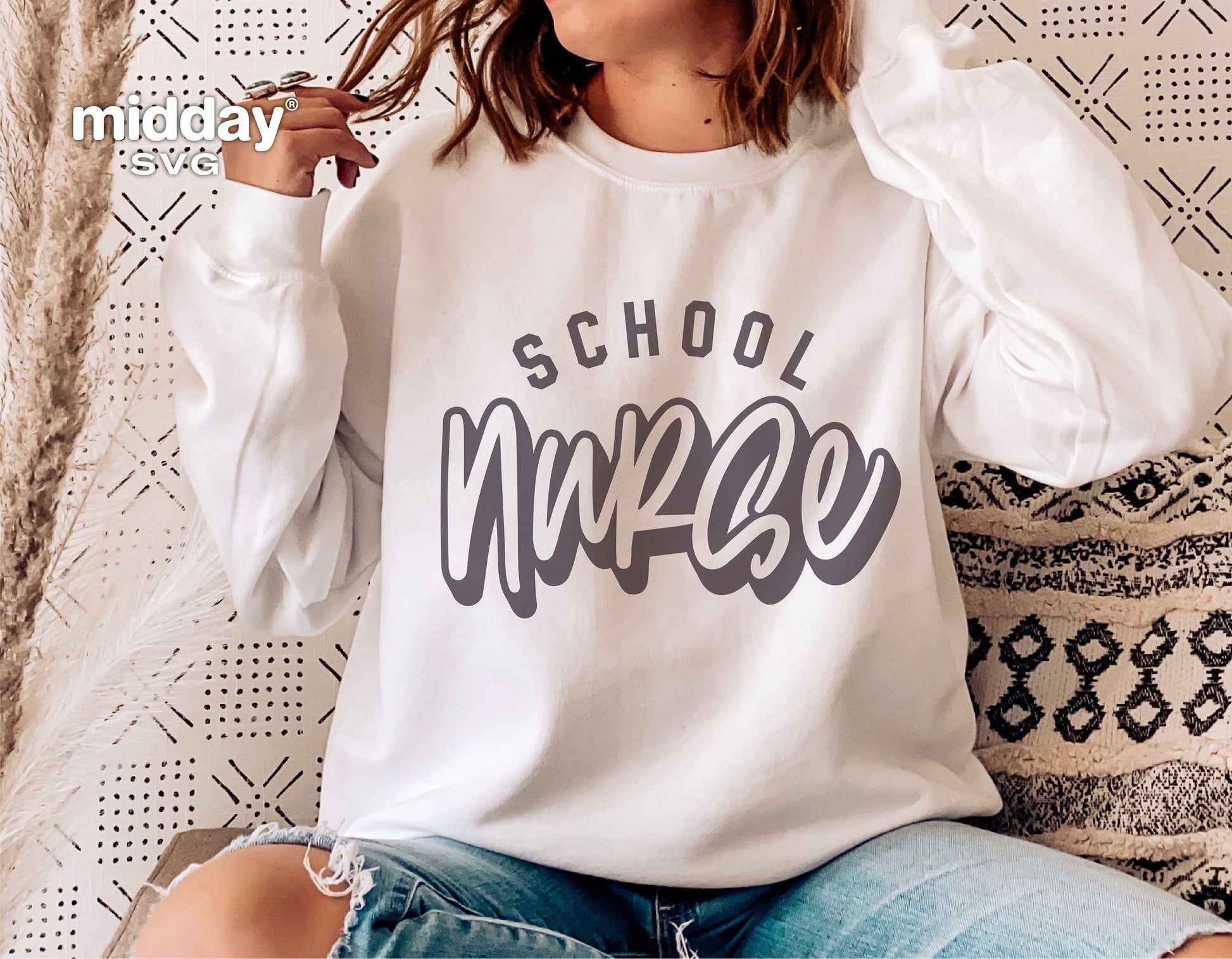 Person wearing a white sweatshirt with 'School Nurse' design in gray, sitting against a patterned background, showcasing casual style.