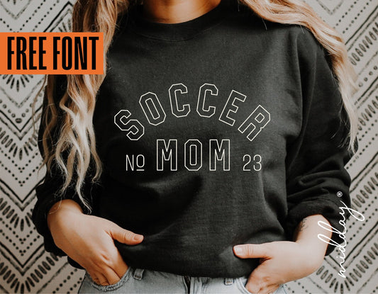 Woman wearing black 'Soccer Mom' sweatshirt with free font offer, perfect for soccer mom gifts, displayed against patterned wall.