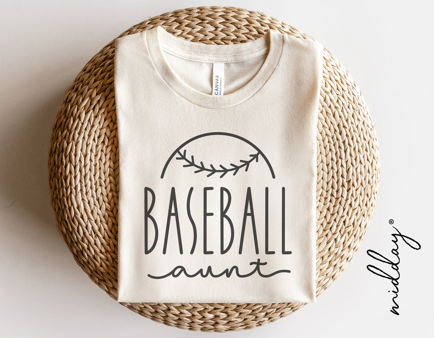 Baseball Aunt shirt design with black text 'Baseball Aunt' and baseball graphic on cream fabric, styled on round woven mat.