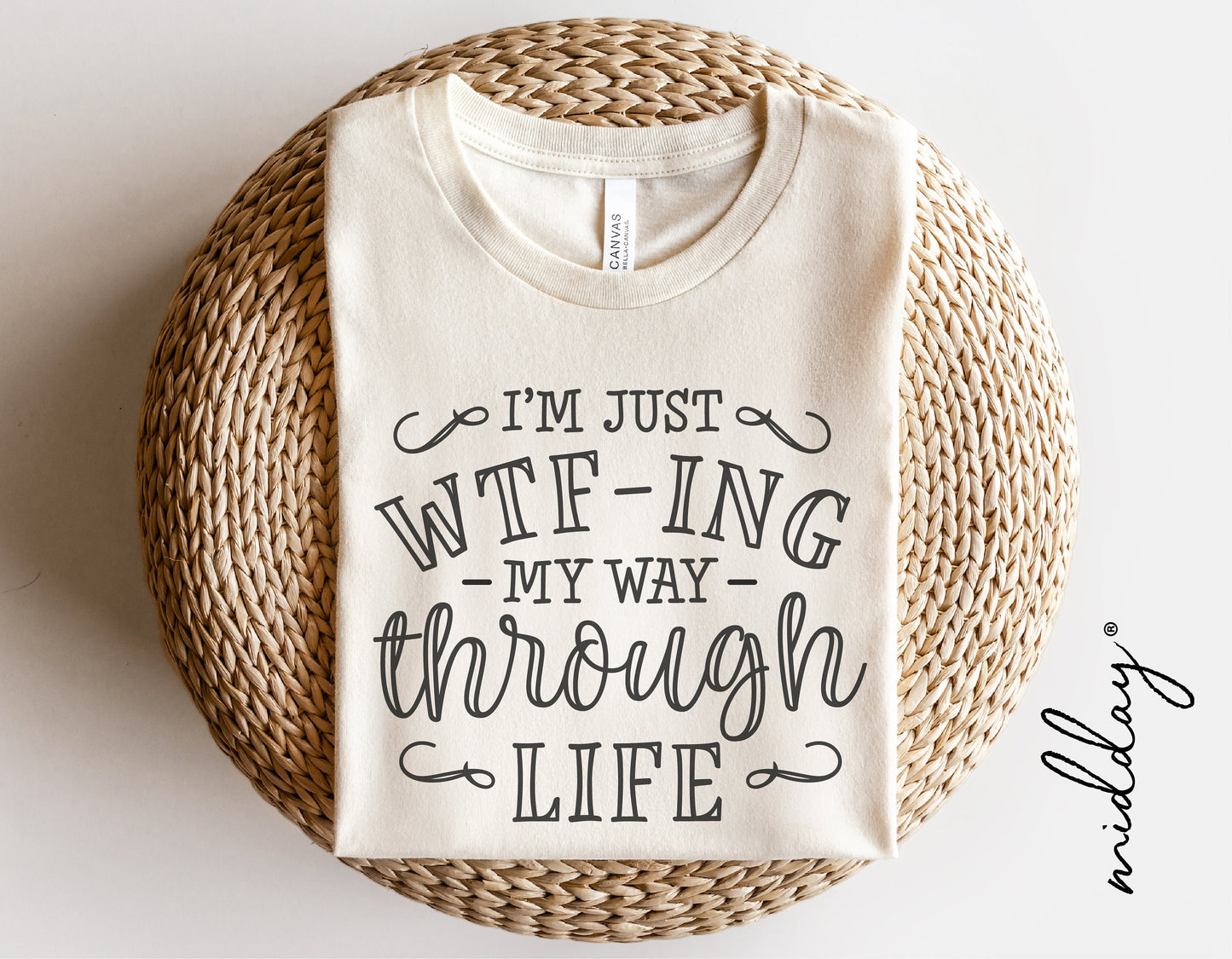 Folded t-shirt with "I'm Just WTF-ing My Way Through Life" printed on it, placed on a woven surface.