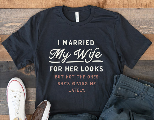 Black t-shirt with "I Married My Wife for Her Looks" funny phrase, displayed with jeans and white sneakers on wooden background.